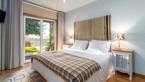 Een slaapkamer met een groot bed, een raam met gordijnen, een nachtkastje met lamp en uitzicht op groen.