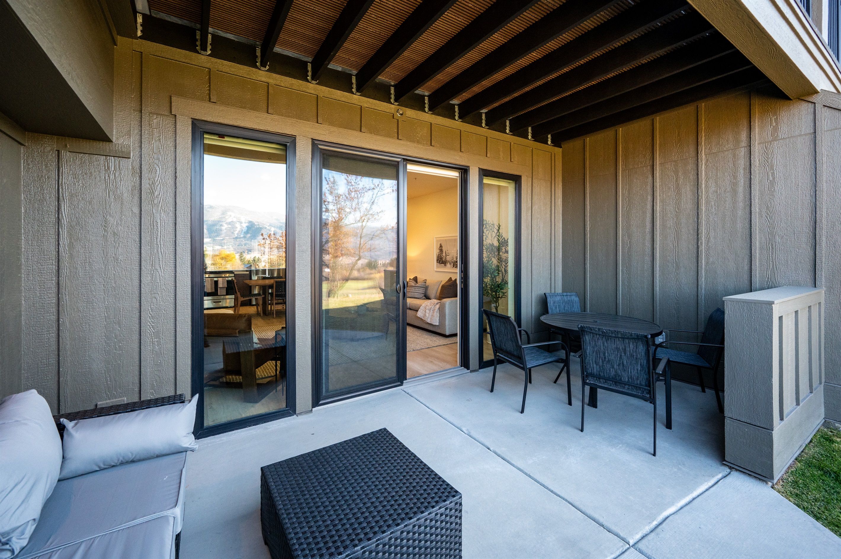 A covered patio area with a sliding glass door leading to an indoor living space, a table with chairs, and a cushioned seating area.