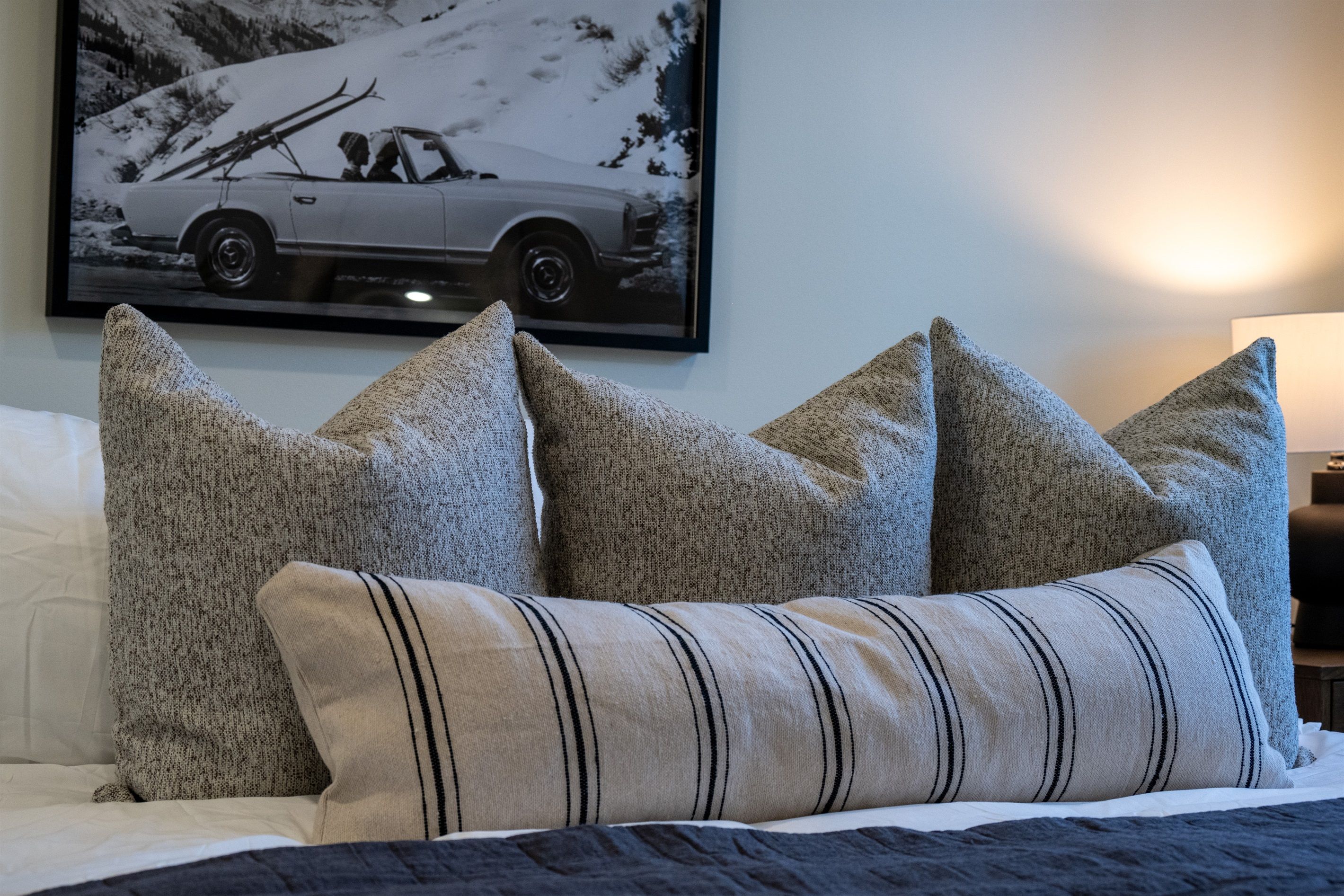 A bed with four pillows, a framed picture of a car on the wall, and a lamp on the side.