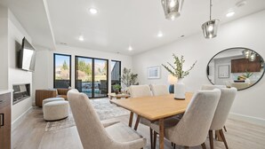 A modern dining area with a wooden table and upholstered chairs, a fireplace, and a large window with a view of the outdoors. - Powder & Golf Pt B106 | Powder Mtn Ski Retreat (Eden)