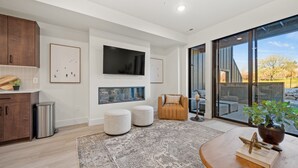 A modern living room with a large TV, ottomans, a sofa, and a view of the outdoors through large windows. - Powder & Golf Pt B106 | Powder Mtn Ski Retreat (Eden)