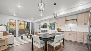 A modern kitchen with wooden cabinets, a dining area with a black table and white chairs, and a large window with a view of a park. - Escape to Eden Pt C103|Ski Getaway |Fireplace (Eden)