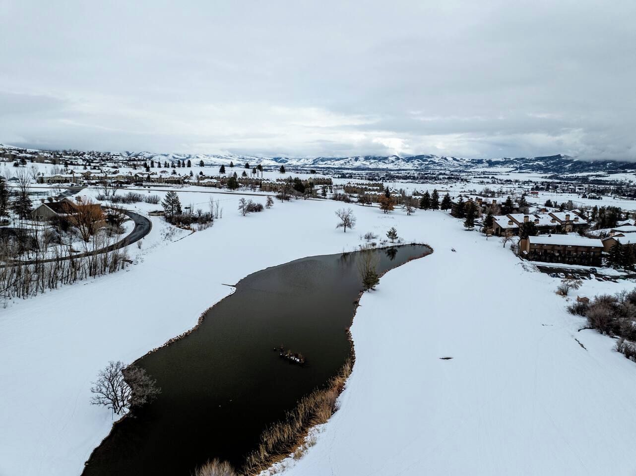 A snow-covered landscape with a river, houses, and mountains in the background.