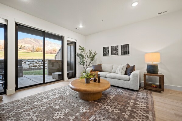 A modern living room with a round wooden coffee table, a grey sofa, and a large sliding glass door leading to an outdoor patio. - Snowbasin Vista Pt C106 | Ski Retreat | Mtn Views (Eden)