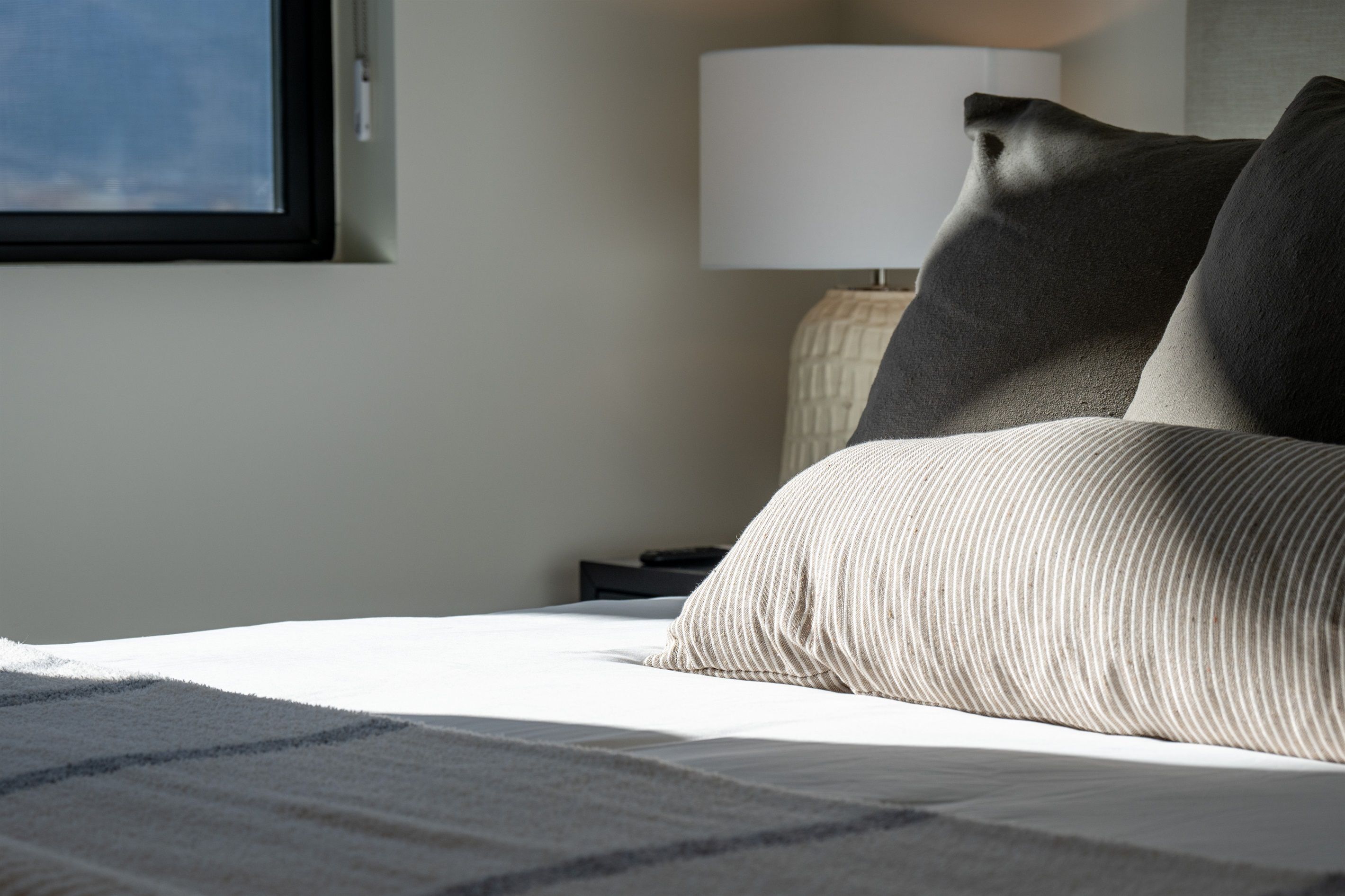 A neatly made bed with a striped blanket and two pillows, a bedside lamp, and a window with a view of the sky.
