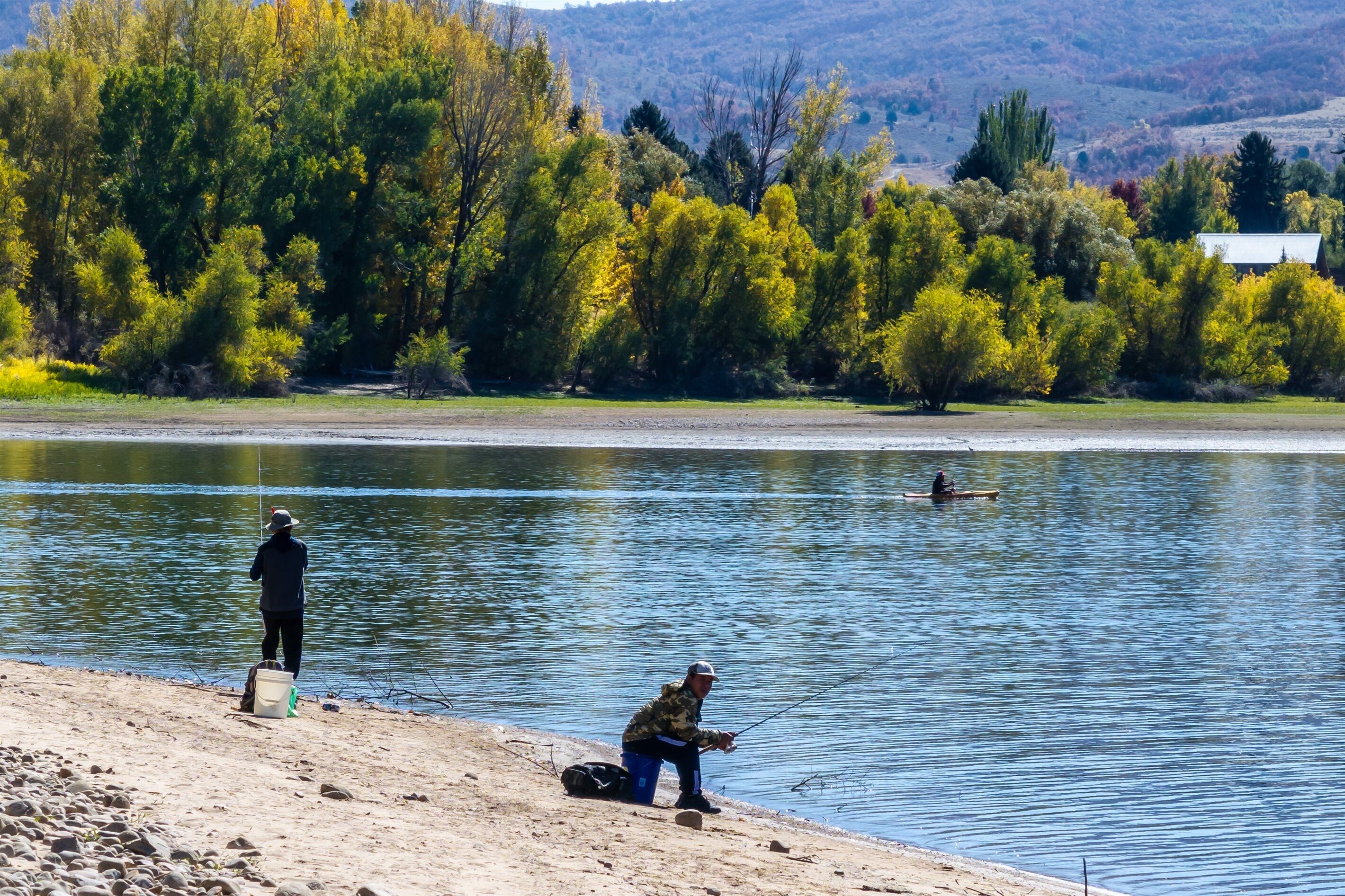 A lakeside scene with two people fishing, a boat, and a house in the background.