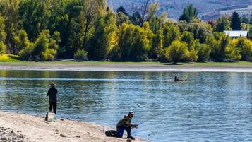 A lakeside scene with two people fishing, a boat, and a house in the background.