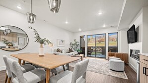 Een moderne woonkamer met een houten eettafel, gestoffeerde grijze stoelen, een grote spiegel en uitzicht op een bergachtig landschap door glazen deuren.