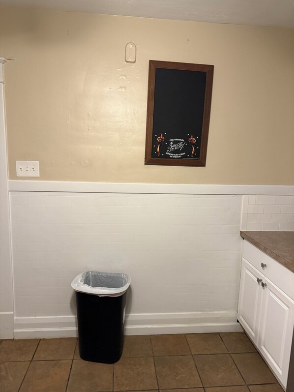 A kitchen with white cabinets, a black trash bin, and a framed sign on the wall.