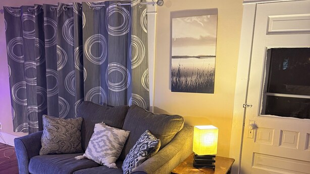 Un salon avec un canapé gris, des coussins à motifs, une table d’appoint avec une lampe et une porte avec une fenêtre.