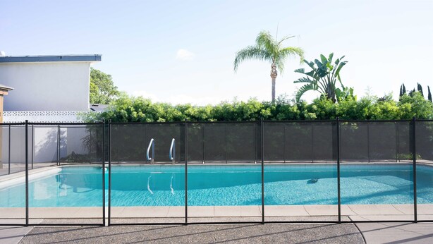 Une piscine entourée d’une clôture de sécurité, bordée de verdure, avec un bâtiment visible au loin.