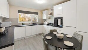 A modern kitchen with white cabinets, a black countertop, and a dining area with a round table and four chairs. - Barnet Oasis near Sky Studios (New Barnet)