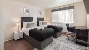 A hotel room with two beds, a desk, a chair, a nightstand, and a window with curtains. - Barnet Oasis near Sky Studios (New Barnet)