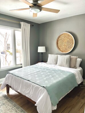 Quarto com cama, ventilador de teto, abajur, espelho redondo fixado na parede e janela com cortinas.