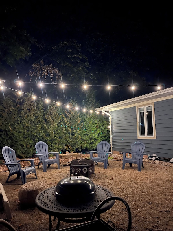 Uma área de pátio ao ar livre com luzes de corda, cadeiras Adirondack e uma mesa redonda com cobertura decorativa.
