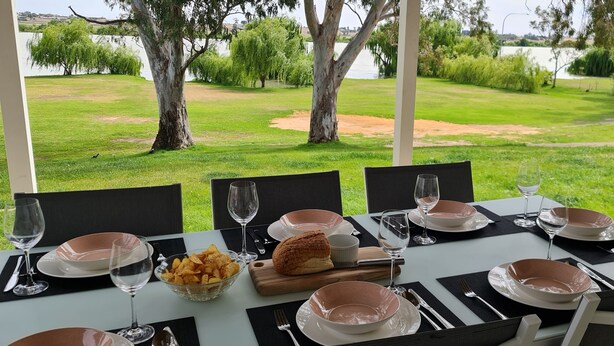An outdoor dining table set with plates, bowls, cutlery, wine glasses, and a basket of chips, overlooking a grassy area with trees and a body of water. - TWIN GUMS - EXCLUSIVE RIVERFRONT PROPERTY - TAILEM BEND (Tailem Bend)