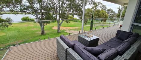 A balcony with a wicker sofa, a glass table, and a view of a grassy area and trees.