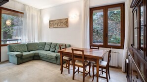 A living room with a green sectional sofa, wooden dining table, and chairs, a painting on the wall, and large windows with curtains. - 'Alassio Holiday' Apartment with Private Garden (Alassio)