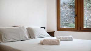 A neatly made bed with white linens and two rolled towels, a wooden nightstand, and a window with a view of a stone wall. - 'Alassio Holiday' Apartment with Private Garden (Alassio)