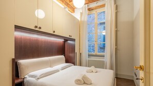 A neatly made bed with pillows and towels, a wooden headboard, a nightstand, and a window with curtains. - San Giorgio A Apartment - Lucca with Wi-Fi (Lucca)