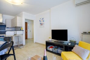 A modern living room with a TV, a dining area with a bar stool, and a kitchen in the background.