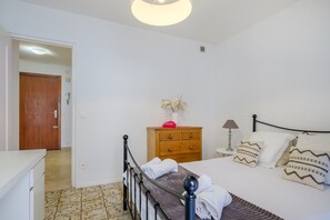 A bedroom with a bed, a wooden dresser, a nightstand, a lamp, and a door leading to another room.