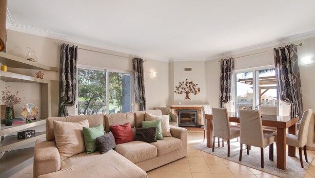 A living room with a beige sofa, a dining table with chairs, a fireplace, and large windows with curtains.