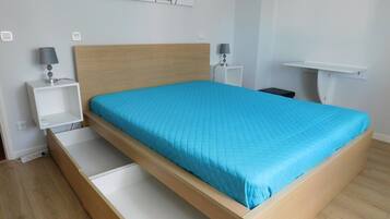 Un dormitorio con estructura de cama de madera, colcha azul, dos mesitas de noche con lámparas, un mueble auxiliar blanco y estante fijado a la pared.