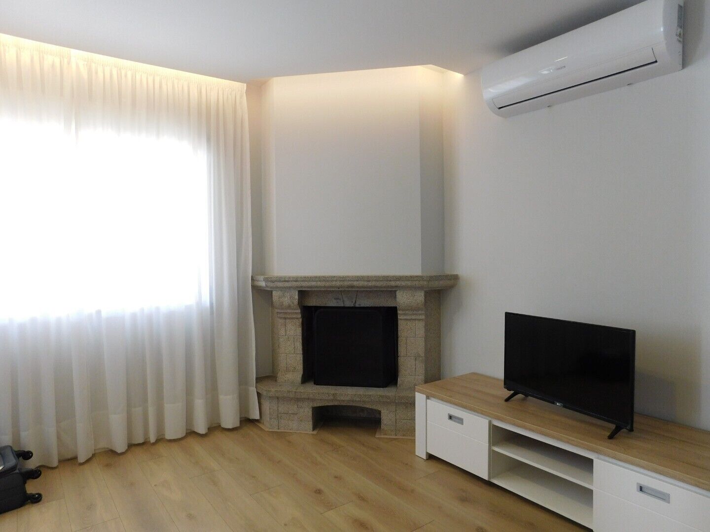 Ein Zimmer mit Kamin, einem Fernseher auf einem Holzständer, einer weißen Klimaanlage an der Wand und einem Fenster mit weißen Vorhängen.