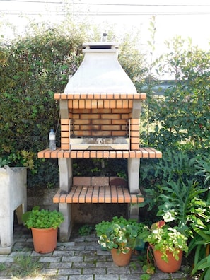 Un horno de ladrillo al aire libre con chimenea, rodeado de follaje y plantas en macetas.