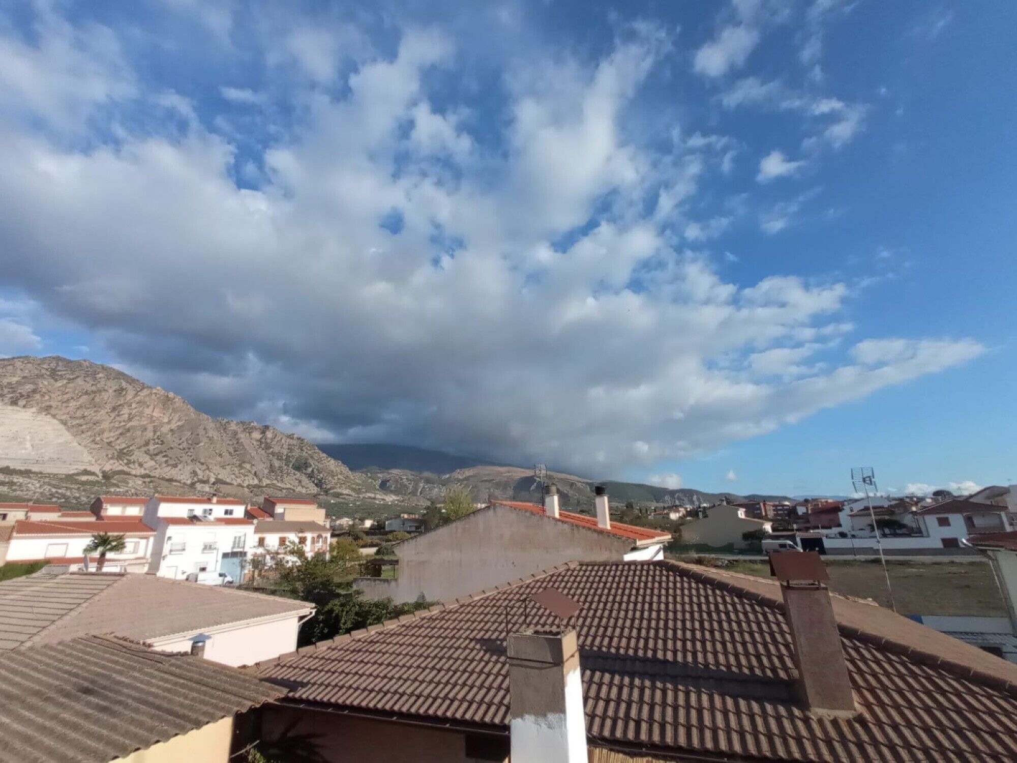 Zona residencial con techos de tejas de terracota, un paisaje montañoso al fondo y cielo parcialmente nublado.