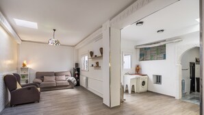 Een moderne woonkamer met een bank, fauteuil en een kleine tafel met een lamp. In de aangrenzende kamer staat een wasmachine en een radiator.