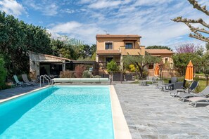 A swimming pool with lounge chairs and umbrellas, surrounded by a patio area and a house in the background.