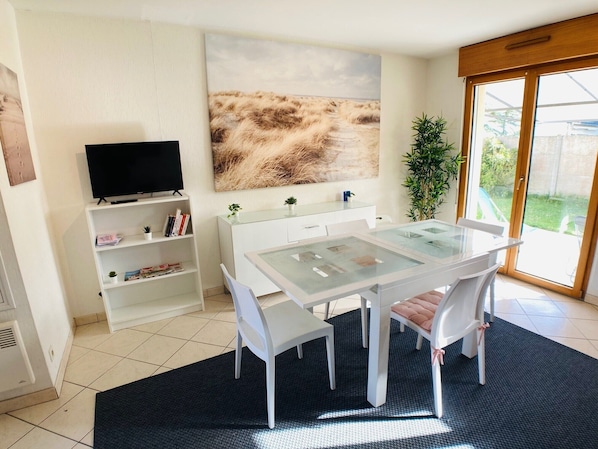 A modern dining area with a glass table, white chairs, a black rug, a bookshelf, a TV, and a large painting.