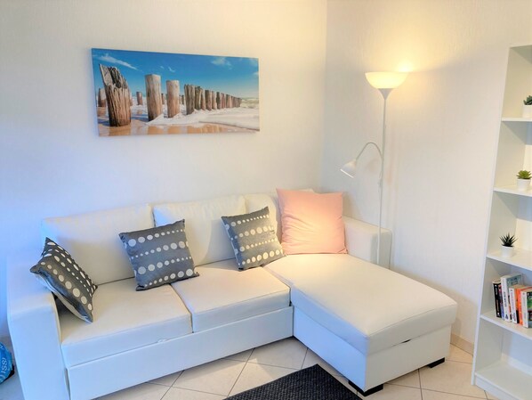 A white sofa with patterned cushions, a painting of a coastal scene, a floor lamp, and a bookshelf with books. - Holiday Home 'Kerann 1min from the Beach with Private Garden', Private Terrace & Wi-Fi (La Turballe)