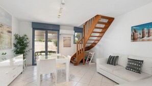 A modern living room with a white sofa, a dining table with chairs, a wooden staircase, and a large window.