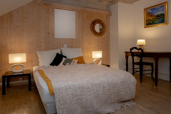 A bedroom with a bed, two bedside tables, a desk, a chair, a round mirror, and a painting on the wall.