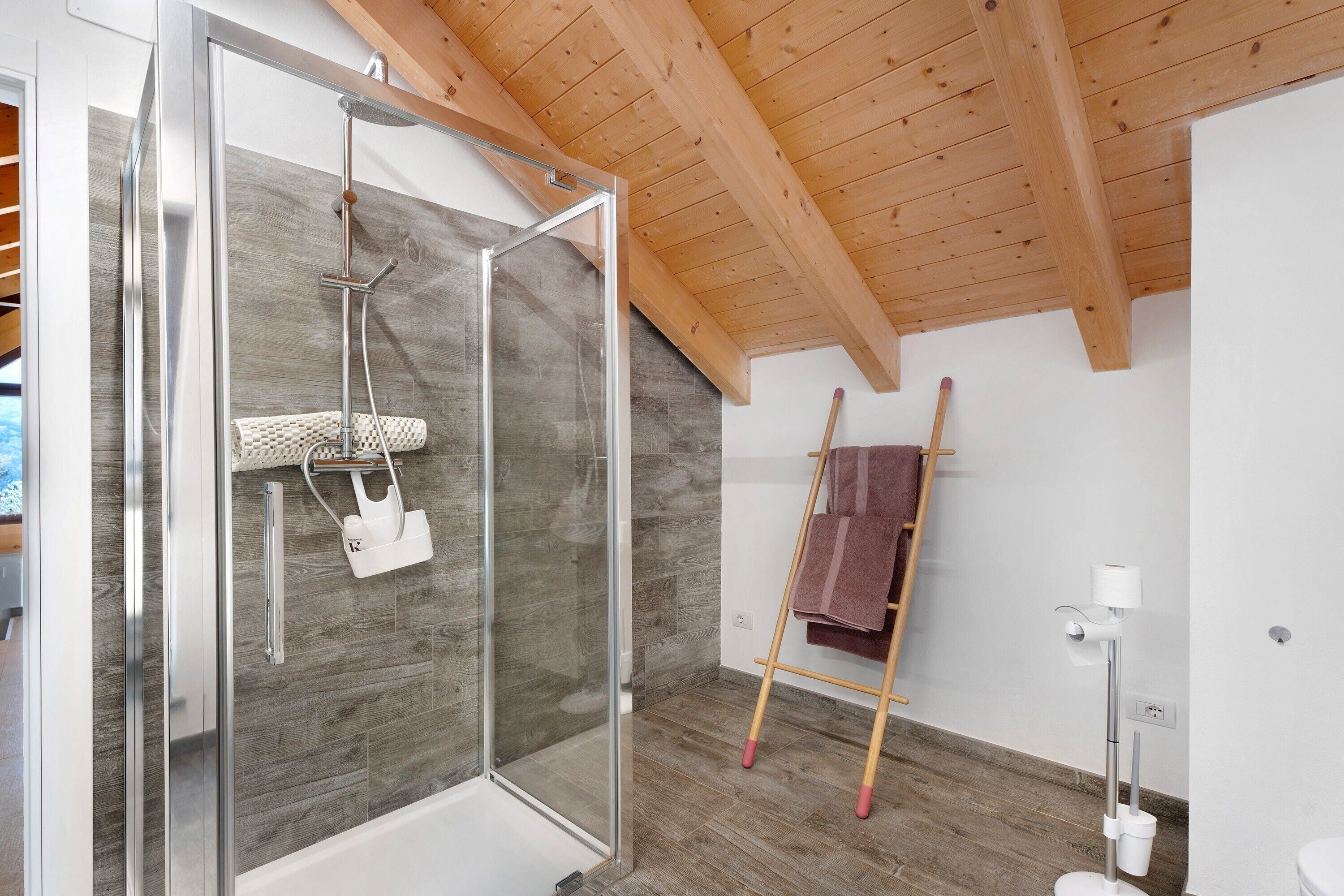 Un baño moderno con cabina de ducha de vidrio, vigas de madera en el techo y una escalera de madera apoyada en la pared.
