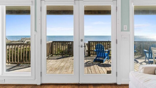Une pièce avec vue sur l’océan, accessible par des portes vitrées, une terrasse en bois à l’extérieur et une chaise bleue sur la terrasse.