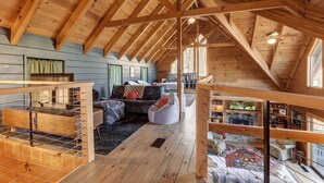 Een ruime zolderkamer met houten balken, een gezellige zithoek met een bank en een relaxfauteuil, en uitzicht op het omliggende landschap.