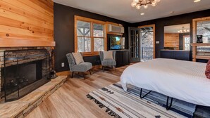 Een slaapkamer met een stenen openhaard, een groot bed, twee stoelen en uitzicht op een bos door een raam.