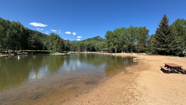 Un entorno tranquilo junto al lago, con una playa arenosa, mesas de picnic y un telón de fondo de árboles y colinas.