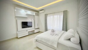 A modern bedroom with a flat-screen TV mounted on a white cabinet, a bed with white bedding, and a window with sheer curtains. - RRR Luxury Villas (Cisarua)