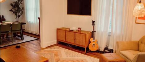 A living room with a wooden coffee table, a sofa, a TV mounted on the wall, and a guitar leaning against the wall.