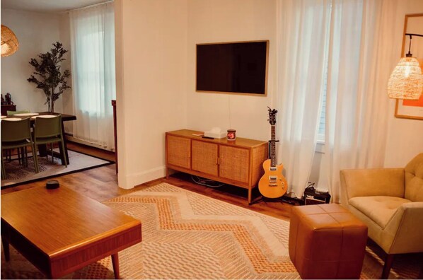 A living room with a wooden coffee table, a sofa, a TV mounted on the wall, and a guitar leaning against the wall.