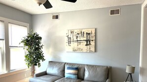 Una sala de estar con un sofá gris, una planta en maceta, un cuadro de un puente y un ventilador de techo.