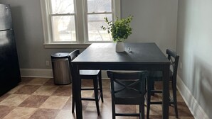 Una cocina con una mesa de comedor, sillas, un refrigerador, un bote de basura y una ventana con persianas.