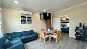 Ein Wohnzimmer mit blauem Sofa, einem Esstisch mit Stühlen, einem an der Wand befestigten Fernseher und einer durchgehende Tür sichtbaren Küchenzeile.