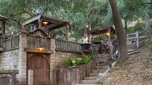 A rustic stone building with a wooden door, a staircase leading up, and a wooden fence.