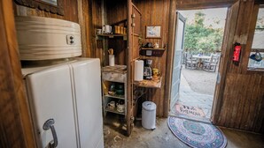 A rustic wooden interior with a white refrigerator, shelves with kitchenware, and a door leading to an outdoor seating area.
